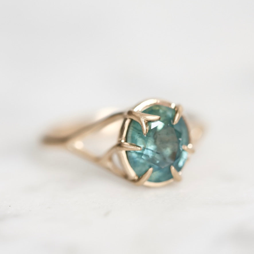A gold ring with a round teal-blue gemstone in a prong setting, photographed close-up on a light marble-like surface.