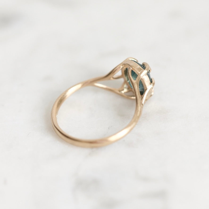 A gold ring with a small oval blue-green gemstone in a prong setting, photographed on a light marble surface.