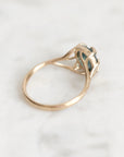 A gold ring with a small oval blue-green gemstone in a prong setting, photographed on a light marble surface.