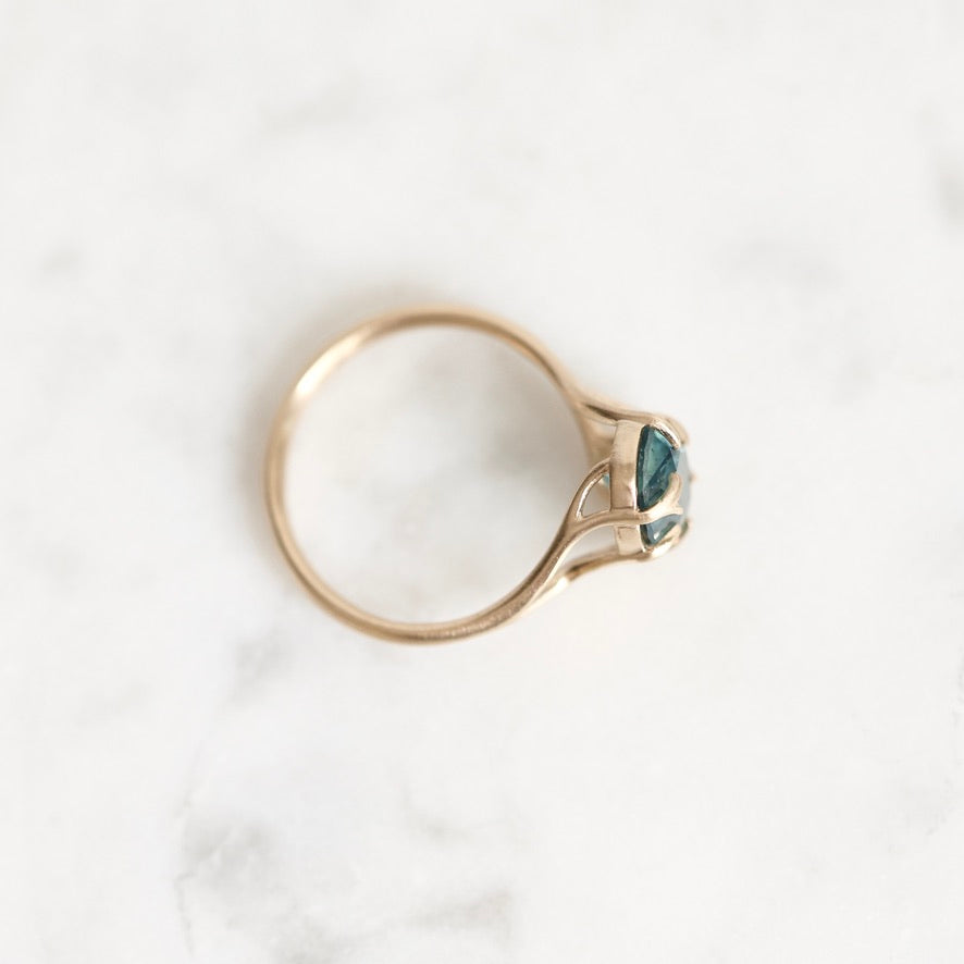 A gold ring with a marquise-cut teal gemstone, photographed from above on a white marble surface.