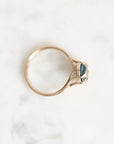 A gold ring with a marquise-cut teal gemstone, photographed from above on a white marble surface.