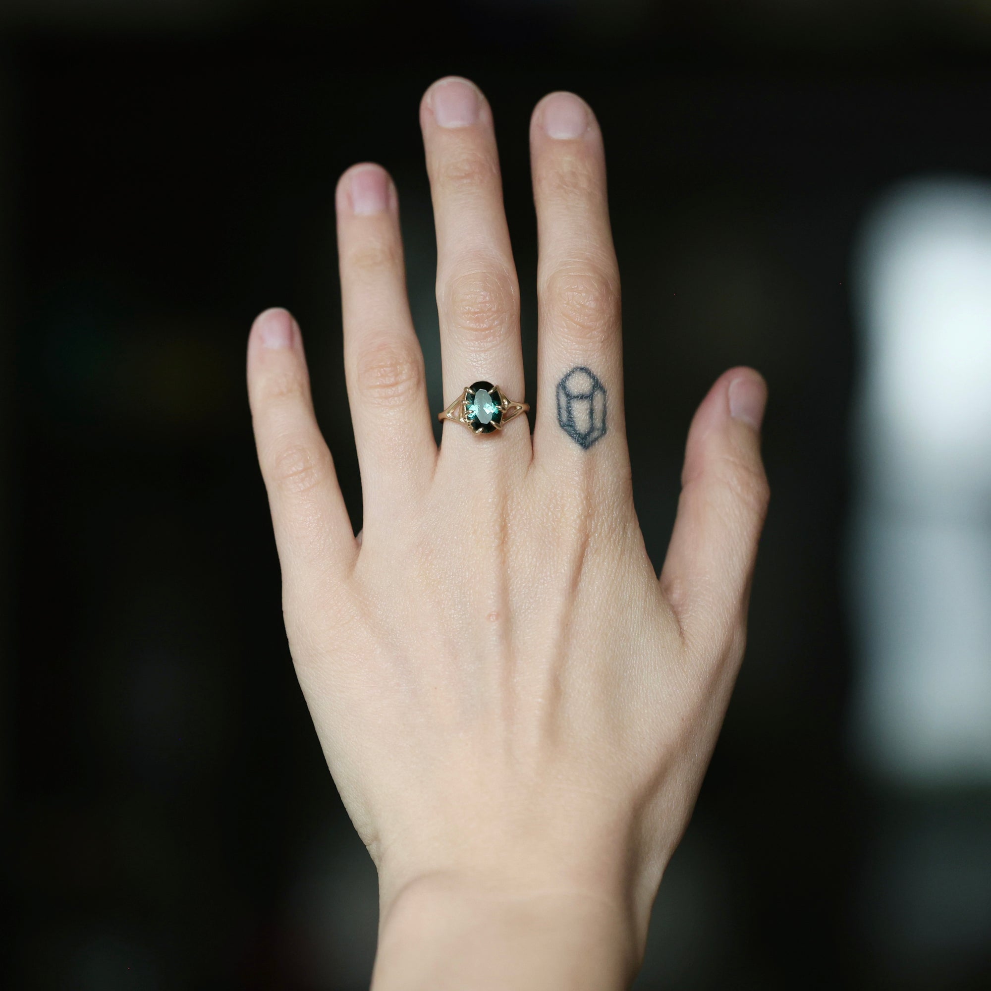 A close-up of a person's hand wearing a ring with a round green gemstone and a small geometric tattoo on the middle finger.
