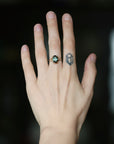 A close-up of a person's hand wearing a ring with a round green gemstone and a small geometric tattoo on the middle finger.