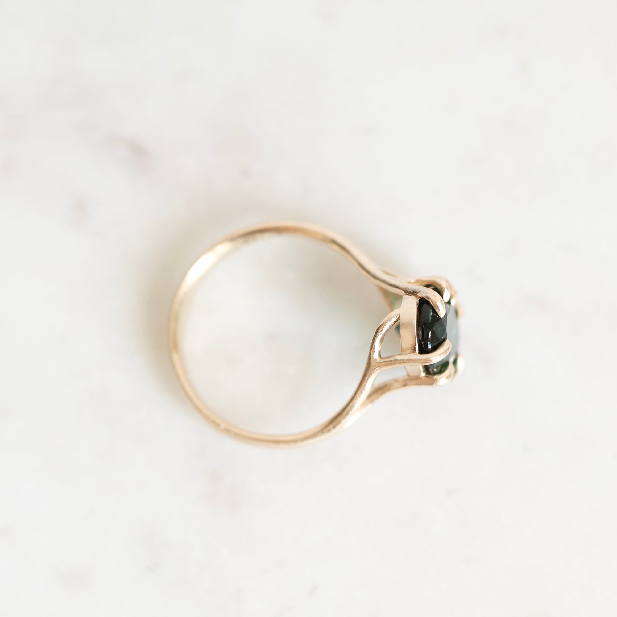 A gold ring with a split shank design and a small dark green rectangular gemstone, photographed on a light marble surface.