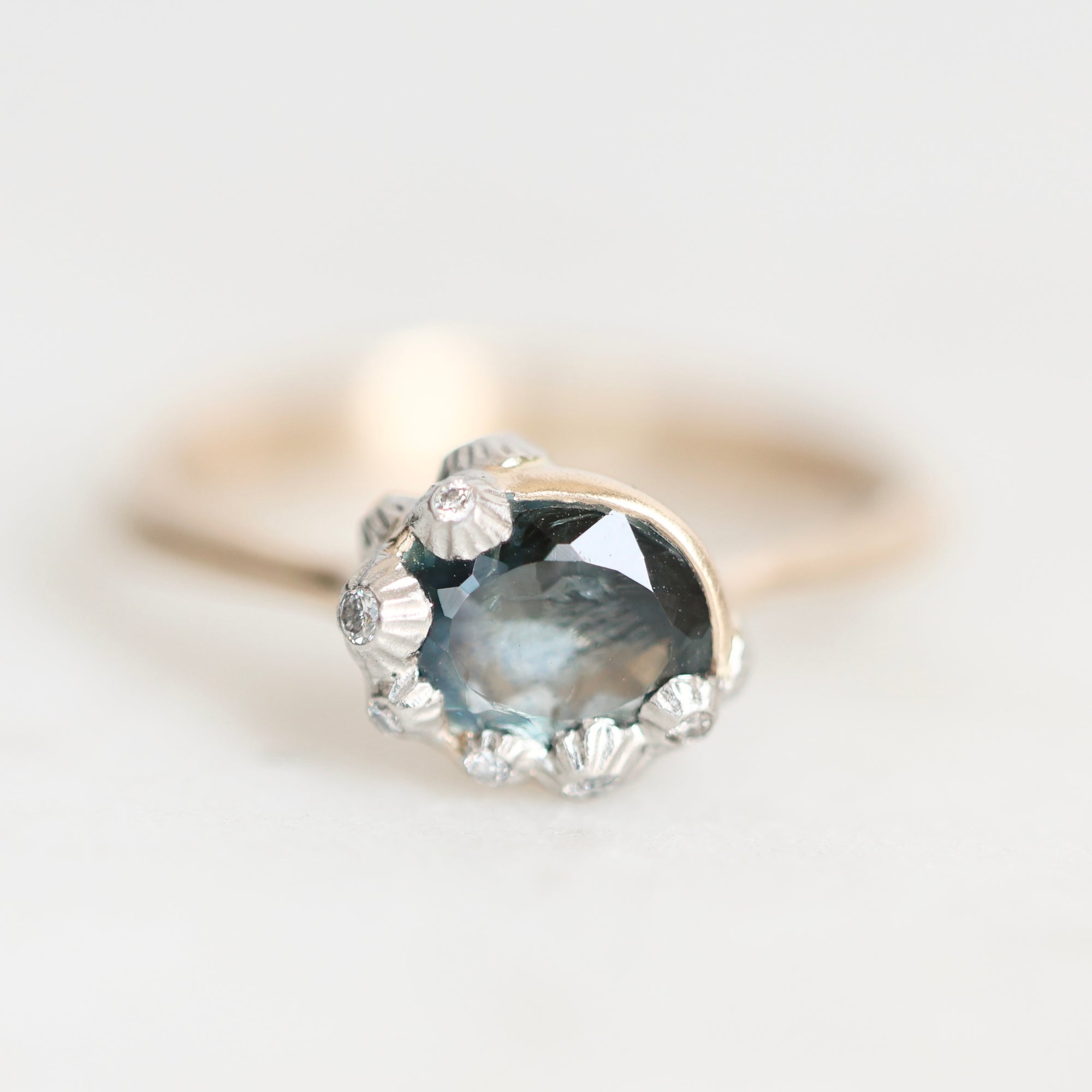 A close-up shot of a ring featuring a large oval blue-gray gemstone in a silver-toned setting, accented with small clear stones, on a soft white background.