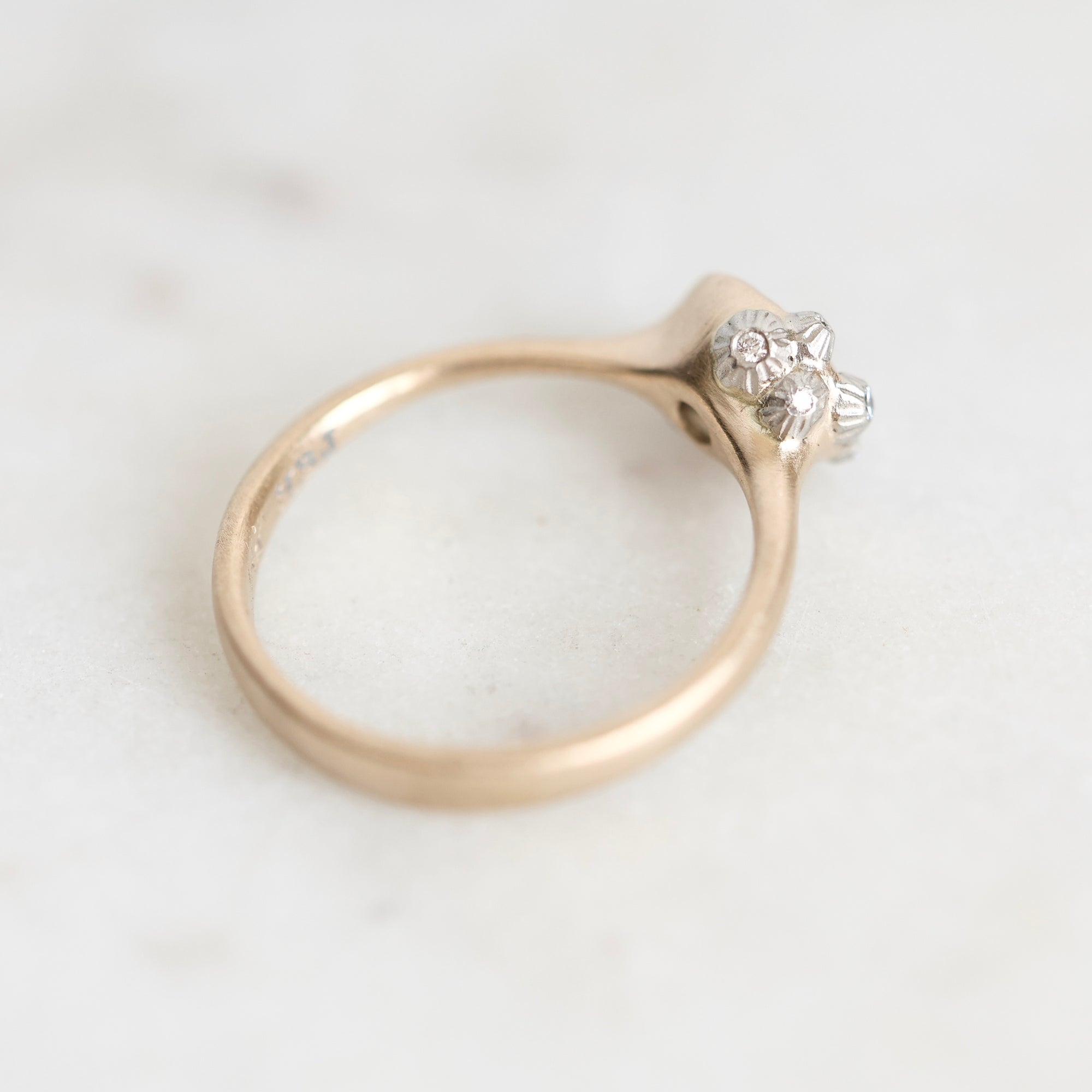 A vintage-style ring with a gold band and a silver-toned floral cluster setting holding small clear stones, photographed on a light marble surface.