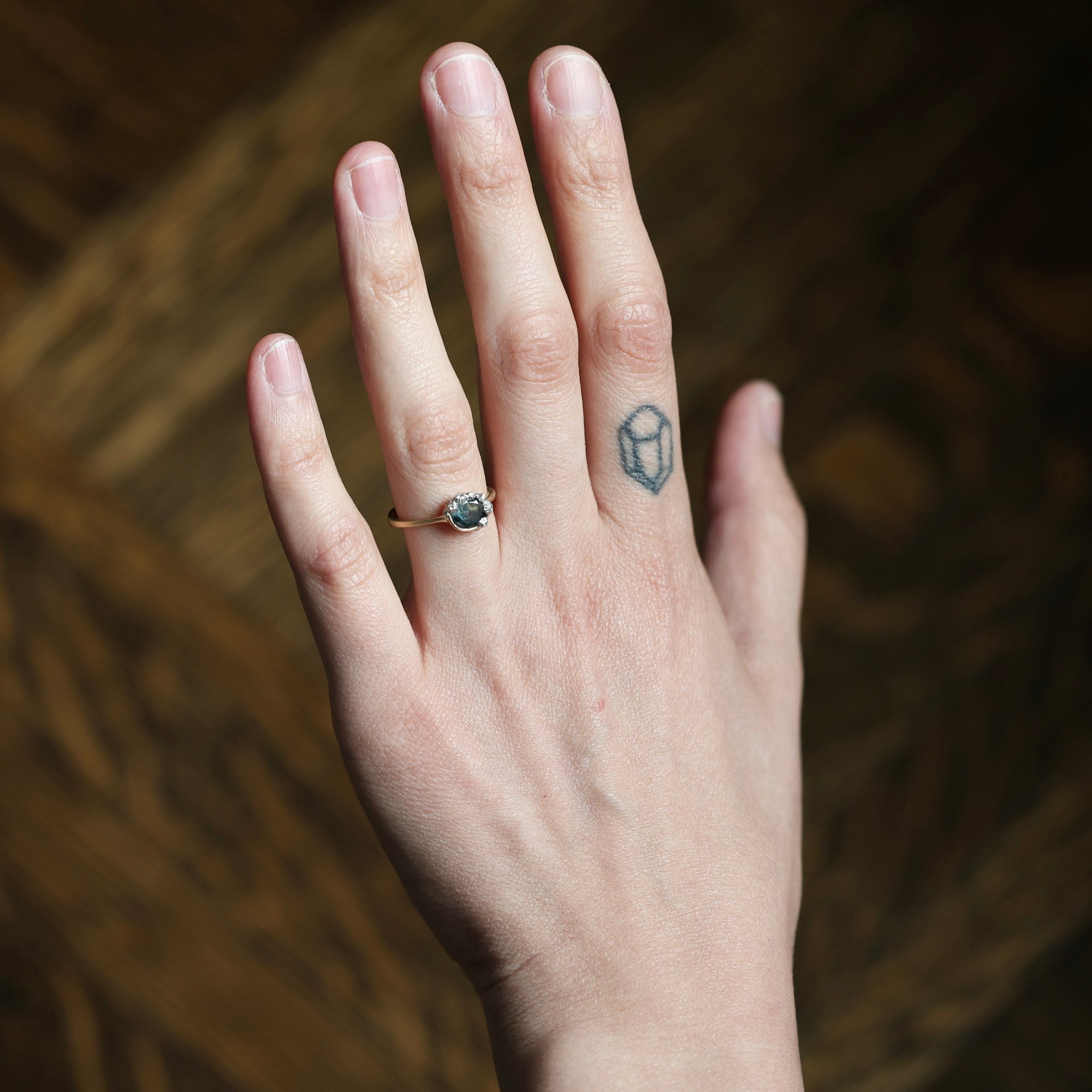 A close-up of a person's hand wearing a delicate rose gold ring with a small oval dark green gemstone, set against a dark brown textured background.
