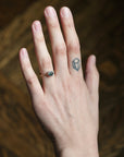 A close-up of a person's hand wearing a delicate rose gold ring with a small oval dark green gemstone, set against a dark brown textured background.