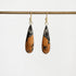 A pair of teardrop-shaped drop earrings with a black and orange marbled pattern, hanging from gold hooks on a horizontal gold rod against a light gray background.