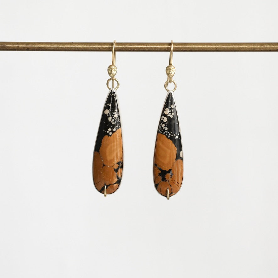 A pair of teardrop-shaped drop earrings with a black and orange marbled pattern, hanging from gold hooks on a horizontal gold rod against a light gray background.