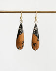 A pair of teardrop-shaped drop earrings with a black and orange marbled pattern, hanging from gold hooks on a horizontal gold rod against a light gray background.