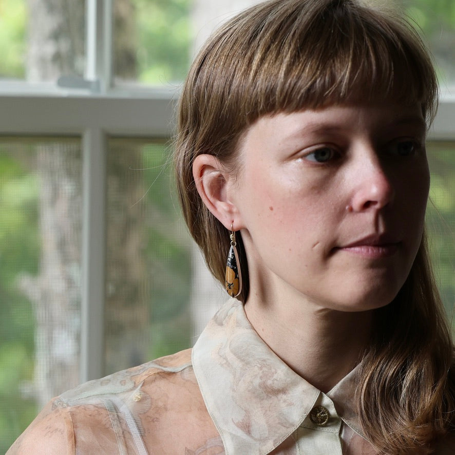 A close-up portrait of a woman wearing a long, teardrop-shaped dangle earring with a gold-toned top and a dark, marbled lower section. She is wearing a sheer, patterned cream blouse in front of a window with soft, natural light.