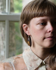 A close-up portrait of a woman wearing a long, teardrop-shaped dangle earring with a gold-toned top and a dark, marbled lower section. She is wearing a sheer, patterned cream blouse in front of a window with soft, natural light.