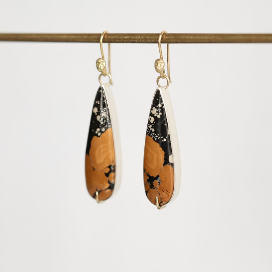 A pair of teardrop-shaped dangle earrings with a black and orange abstract pattern, suspended from gold fishhook wires and hanging from a gold rod against a plain light background.