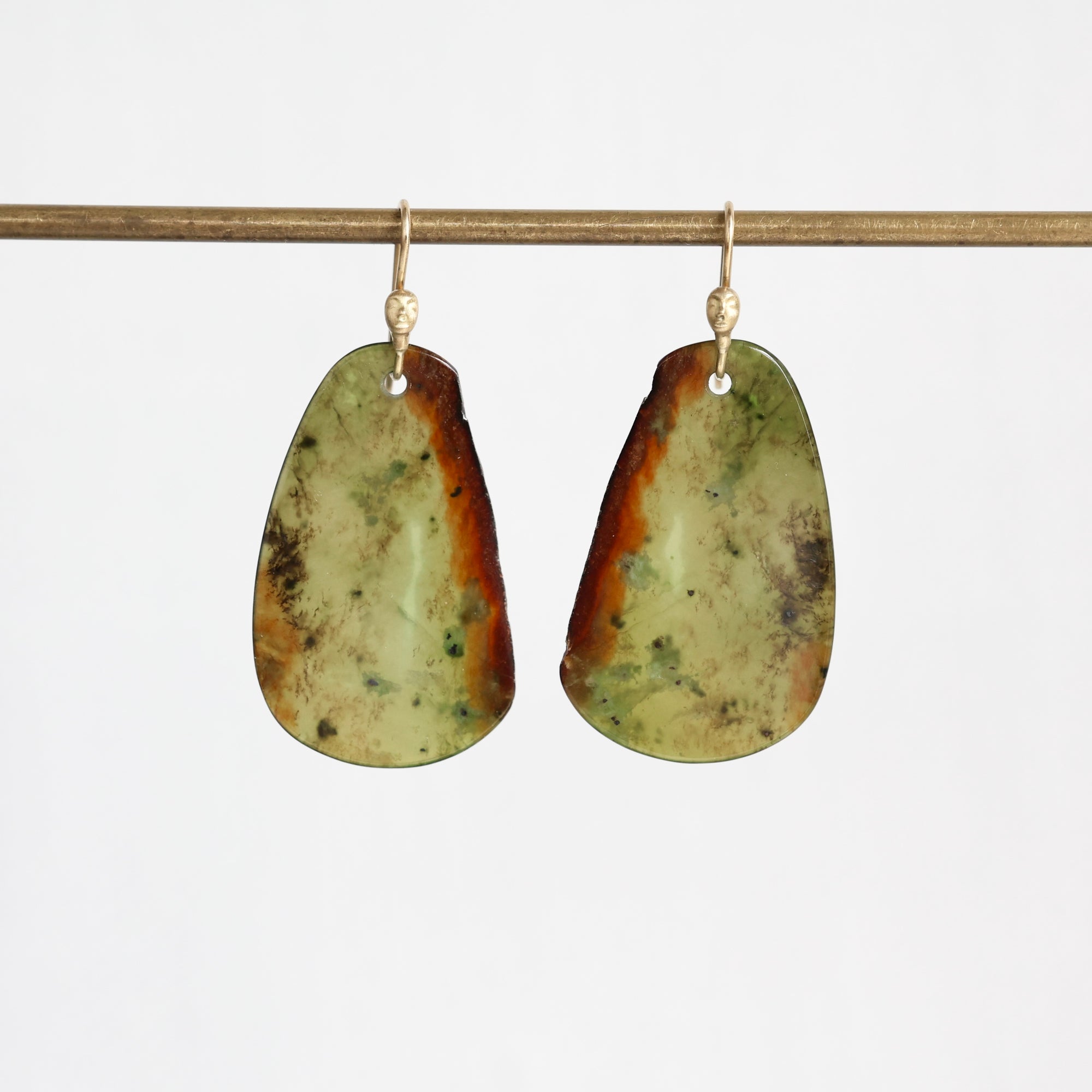 A pair of organic-shaped dangle earrings made from polished green and brown stone, hanging from gold fishhook wires against a plain white background.