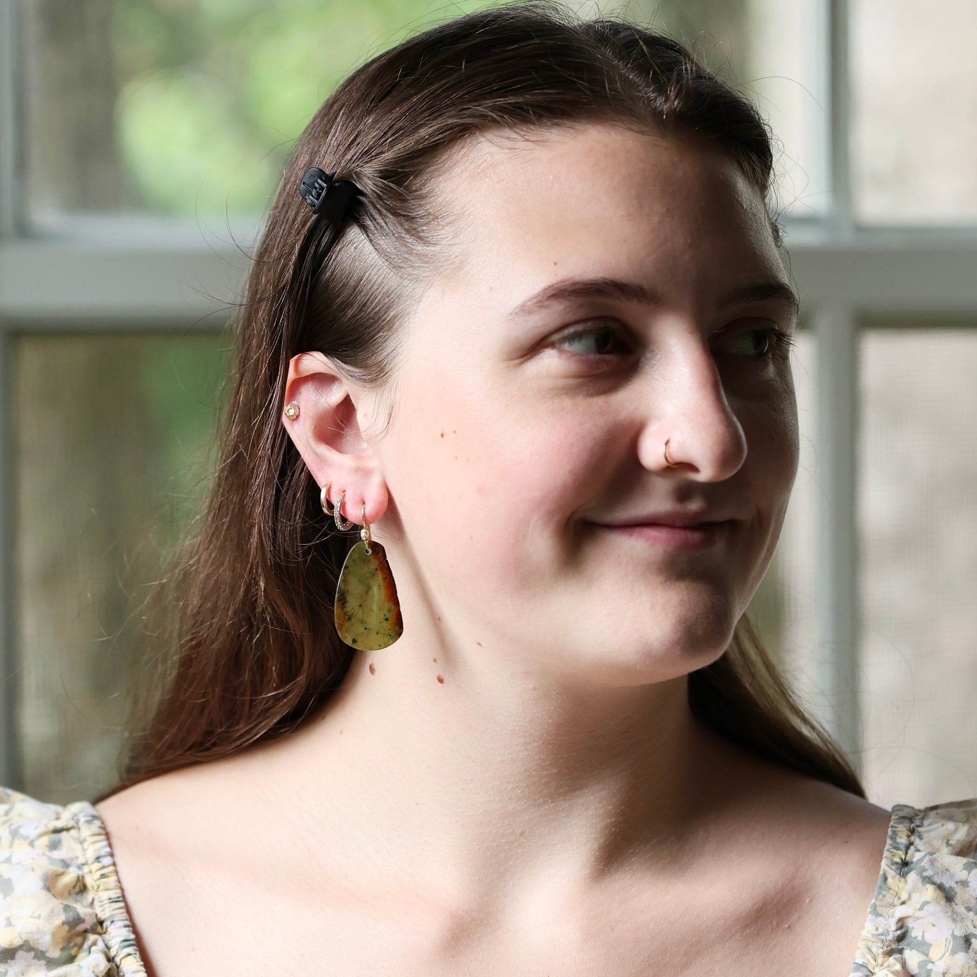A woman with brown hair wearing a black hair clip and a dangling olive-green leaf earring, shown in a close-up portrait against a soft-focus window background.