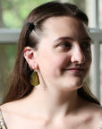 A woman with brown hair wearing a black hair clip and a dangling olive-green leaf earring, shown in a close-up portrait against a soft-focus window background.