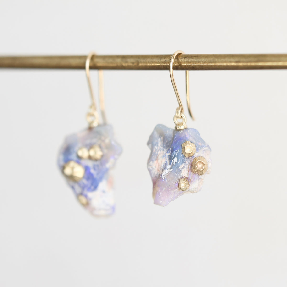 A pair of dangle earrings featuring iridescent blue and white fossilized opal with small gold metallic accents, hanging from gold-toned French hook wires on a gold-colored horizontal rod against a light gray background.