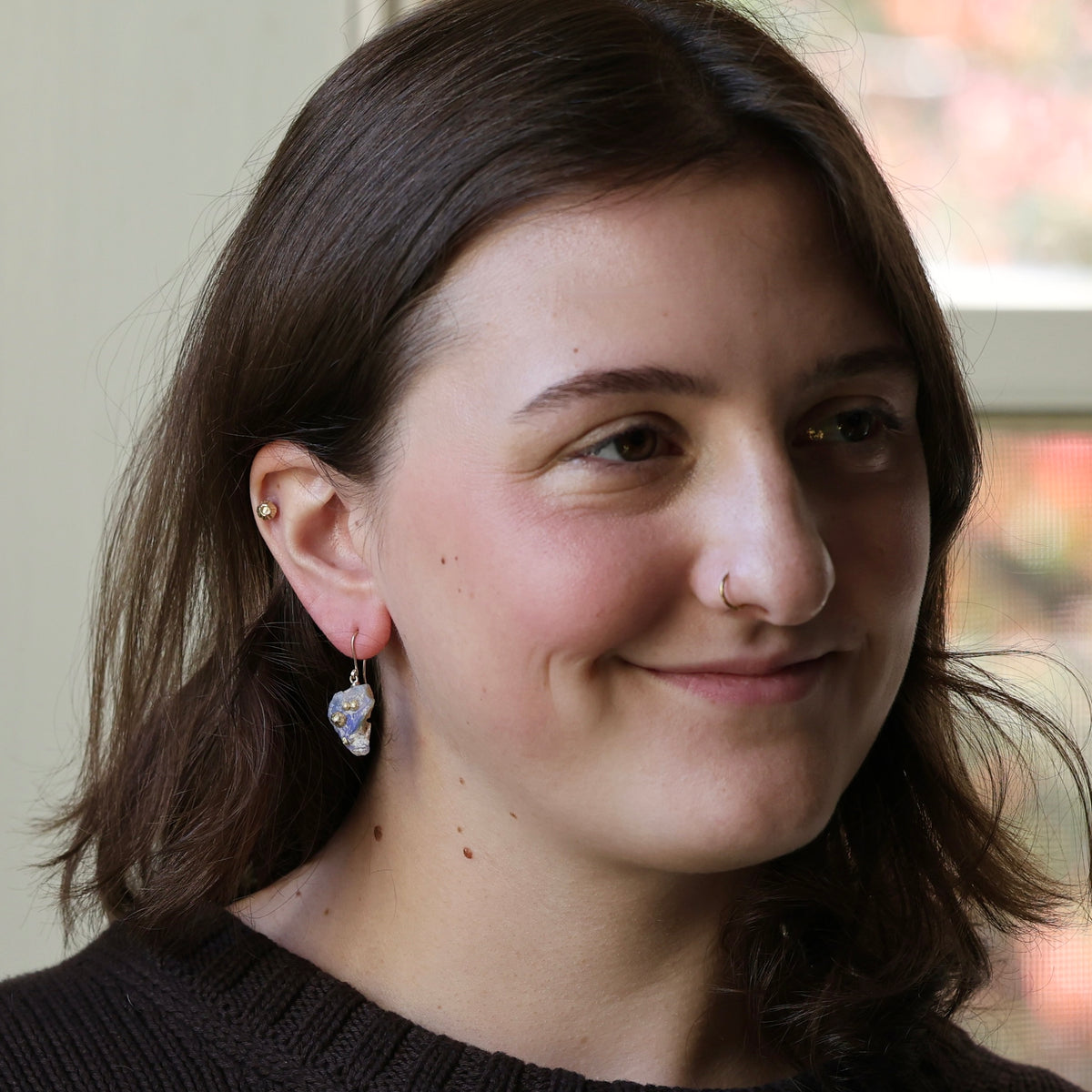 A close-up portrait of a woman with brown hair wearing a silver-toned dangle earring with  stone, set against an indoor background.