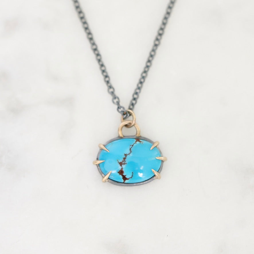 A close-up shot of a necklace featuring an oval turquoise pendant with a gold bezel and prongs, suspended from a dark gray chain on a white marble background.