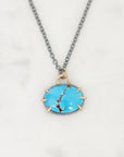 A close-up shot of a necklace featuring an oval turquoise pendant with a gold bezel and prongs, suspended from a dark gray chain on a white marble background.