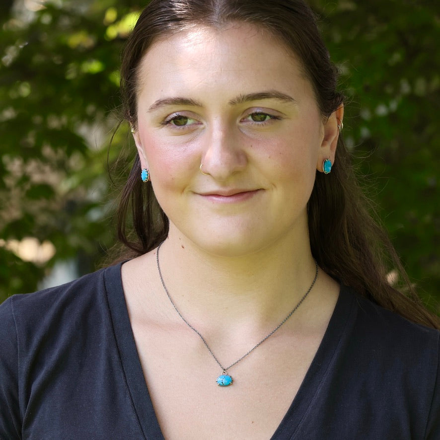 A woman with dark hair wearing a silver necklace with a turquoise pendant and matching turquoise stud earrings, photographed outdoors against a blurred green foliage background.