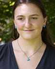 A woman with dark hair wearing a silver necklace with a turquoise pendant and matching turquoise stud earrings, photographed outdoors against a blurred green foliage background.