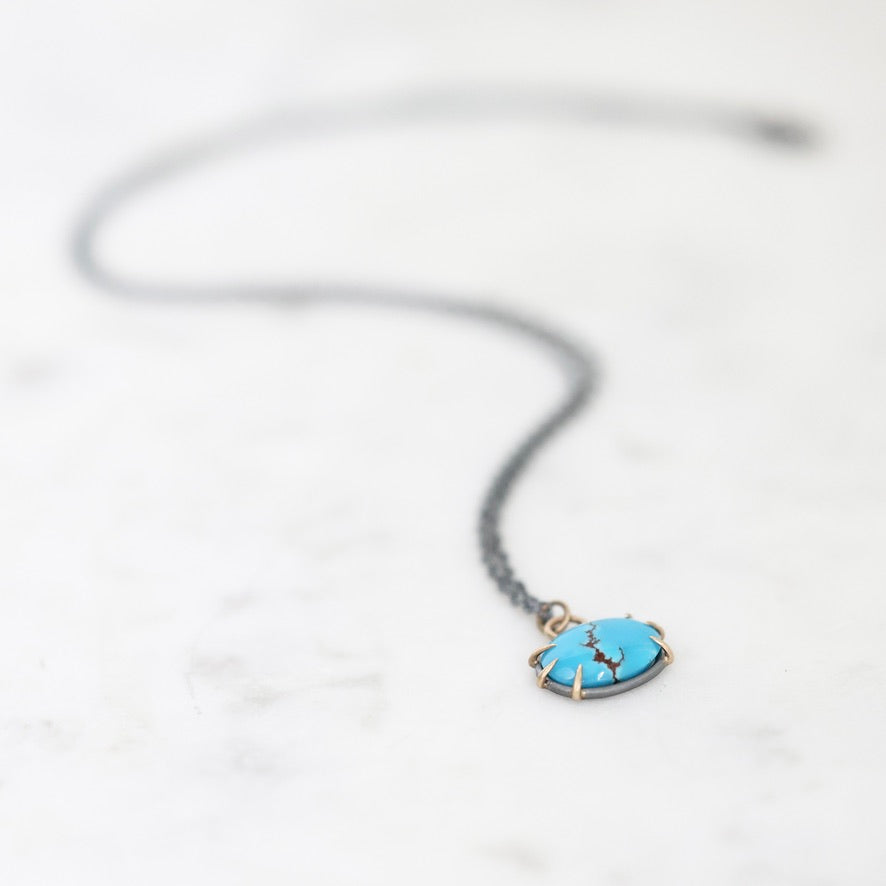 A delicate necklace with a small oval turquoise pendant in a gold-toned bezel setting, suspended from a thin dark chain on a light marble surface.