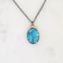 A turquoise stone pendant necklace with a silver-toned chain and gold-toned prong setting, displayed on a white marble surface.