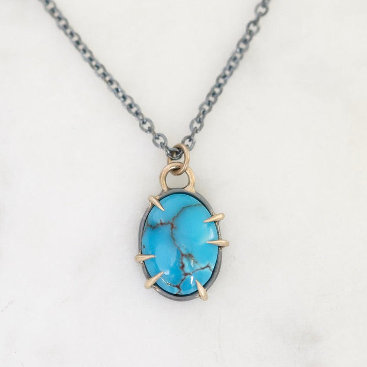 A turquoise stone pendant necklace with a silver-toned chain and gold-toned prong setting, displayed on a white marble surface.