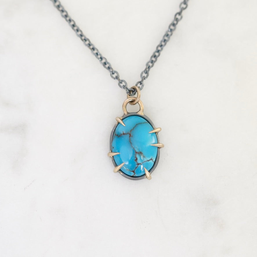 A turquoise stone pendant necklace with a silver-toned chain and gold-toned prong setting, displayed on a white marble surface.