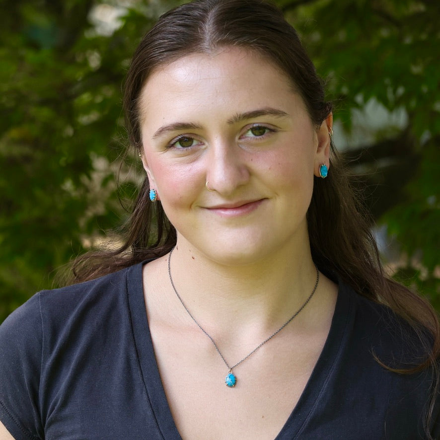 A woman with long brown hair wearing a silver necklace with a turquoise pendant and matching turquoise stud earrings, photographed outdoors against a blurred green foliage background.