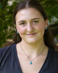 A woman with long brown hair wearing a silver necklace with a turquoise pendant and matching turquoise stud earrings, photographed outdoors against a blurred green foliage background.
