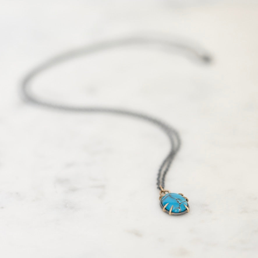 A delicate dark gray chain necklace with a small oval pendant featuring a bright turquoise blue center and gold-toned edges, resting on a light marble surface.