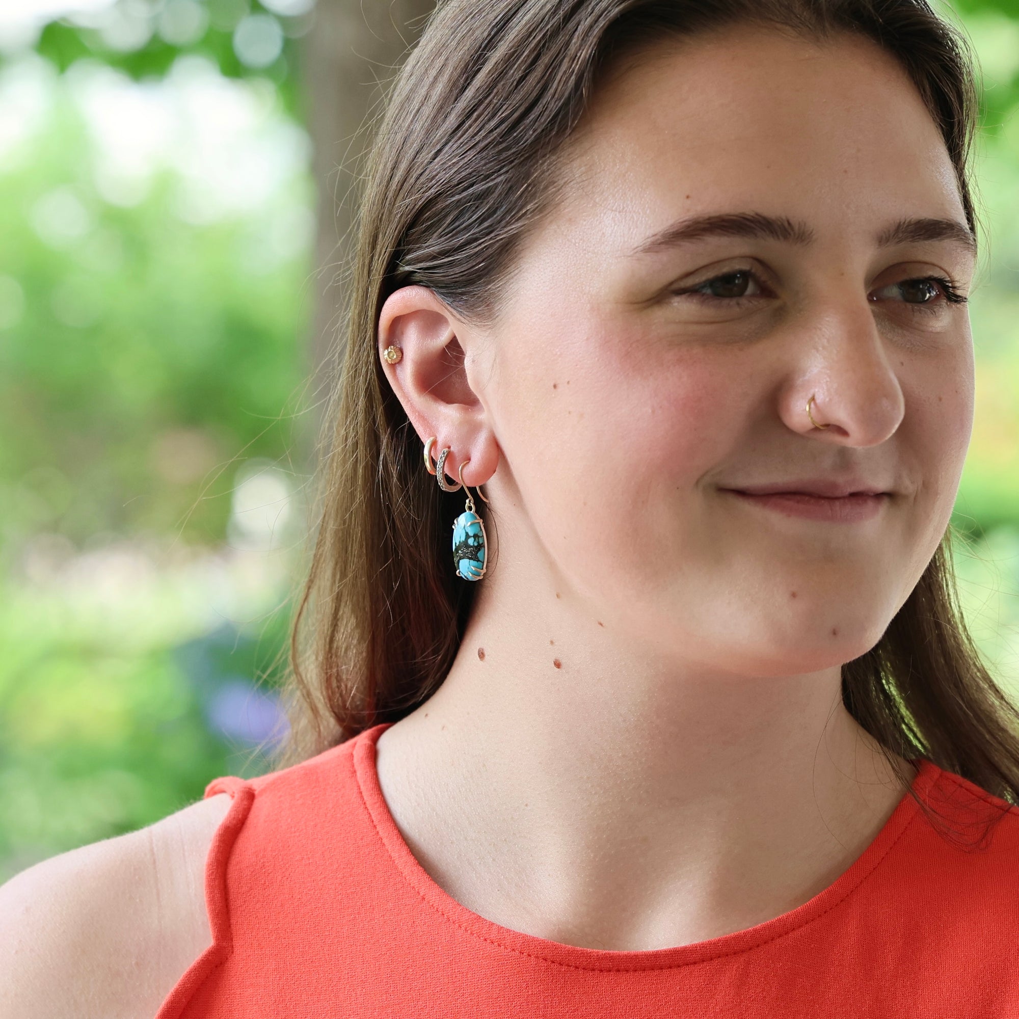 A close-up portrait of a woman wearing a dangling earring with a blue and white patterned stone or bead, photographed outdoors with a blurred green background.