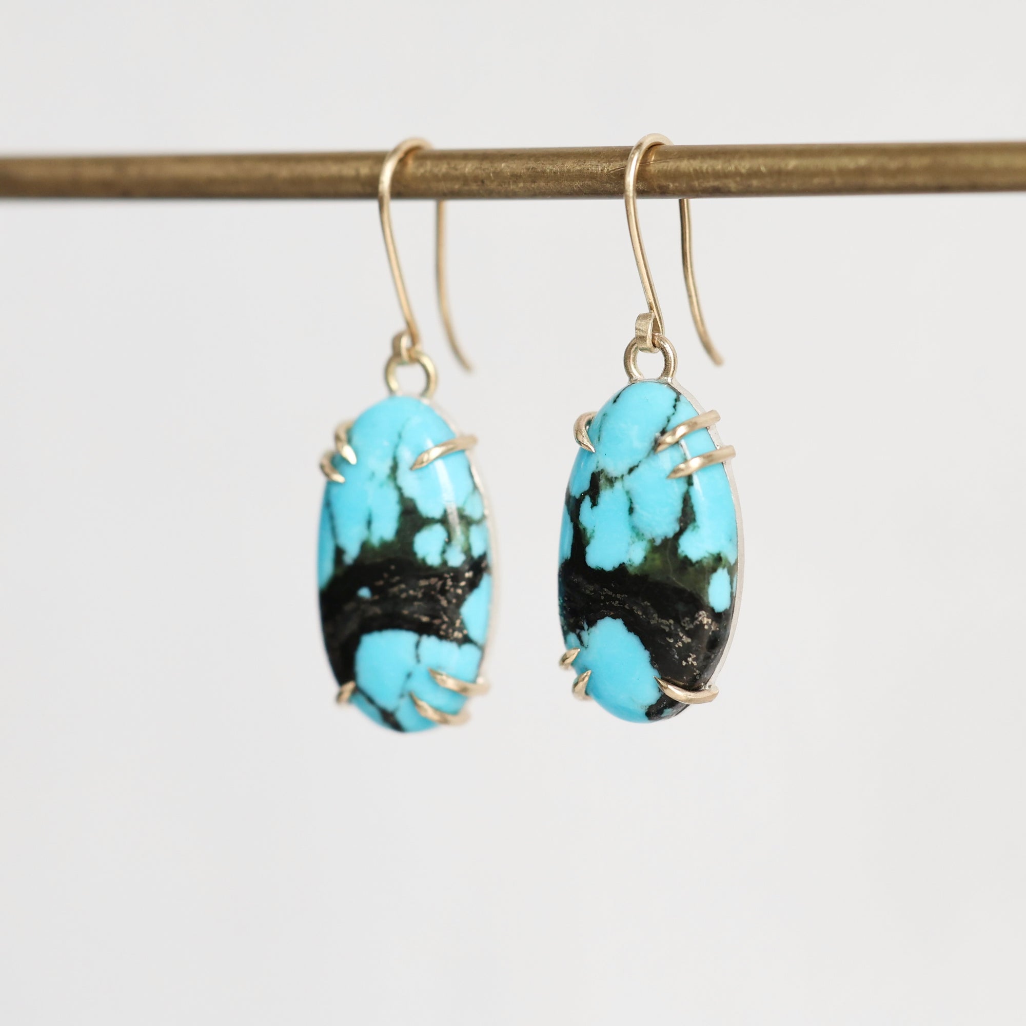 A pair of drop earrings with oval turquoise-blue and black marbled stone pendants in gold wire wrapping and gold fishhook ear wires, hanging from a gold horizontal bar against a light gray background.