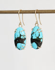 A pair of drop earrings with oval turquoise-blue and black marbled stone pendants in gold wire wrapping and gold fishhook ear wires, hanging from a gold horizontal bar against a light gray background.