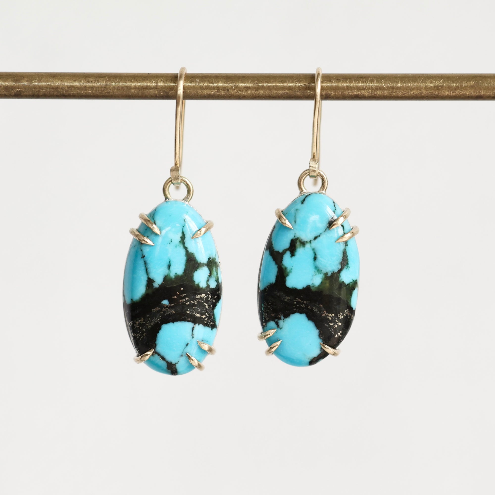 A pair of oval drop earrings with bright turquoise blue and black marbled stones, held in gold-toned prong settings with gold hooks, hanging from a gold rod against a light gray background.
