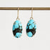A pair of oval drop earrings with bright turquoise blue and black marbled stones, held in gold-toned prong settings with gold hooks, hanging from a gold rod against a light gray background.