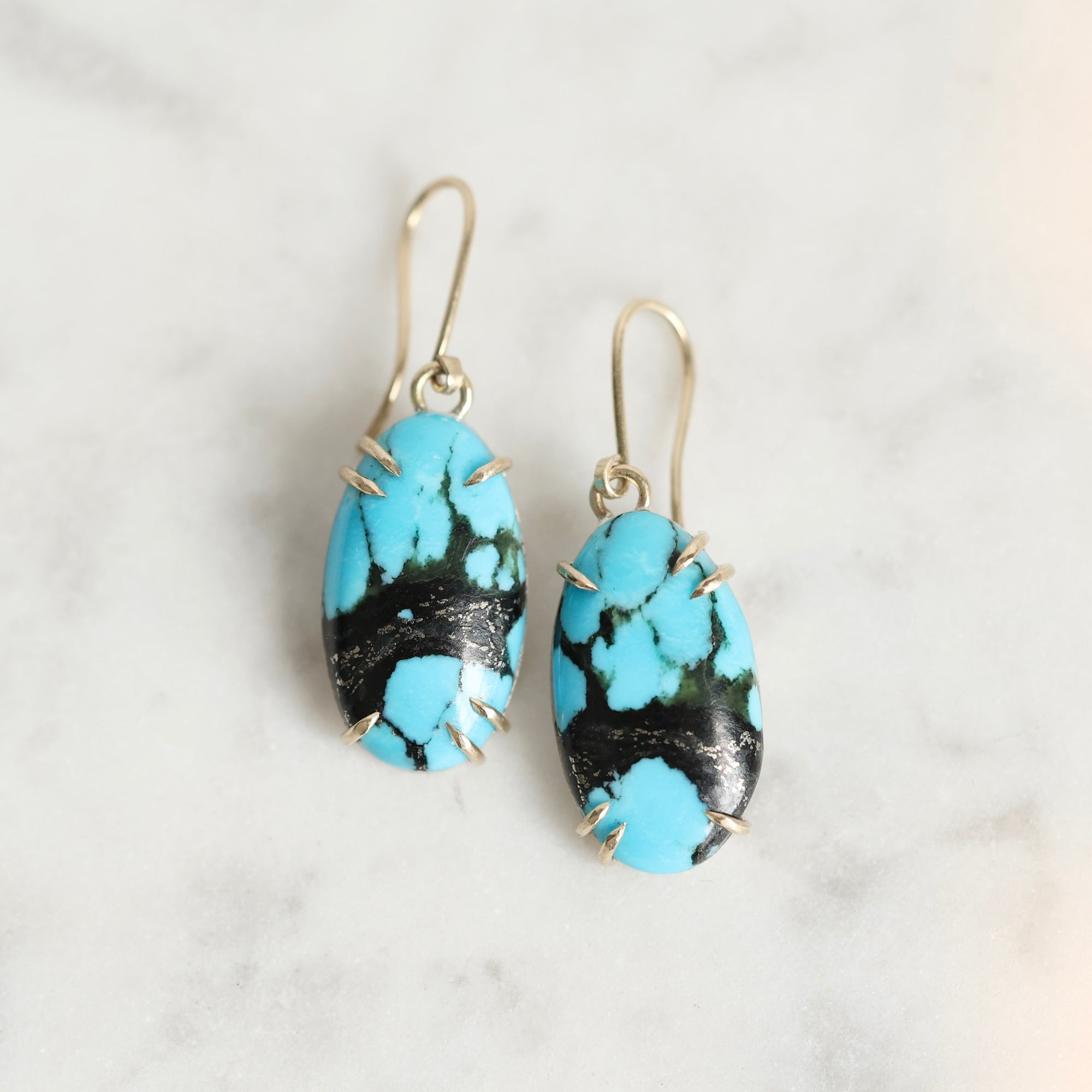 A pair of drop earrings with oval turquoise and black marbled stones in silver wire wrap, hanging from gold-tone fishhook wires on a light marble surface.