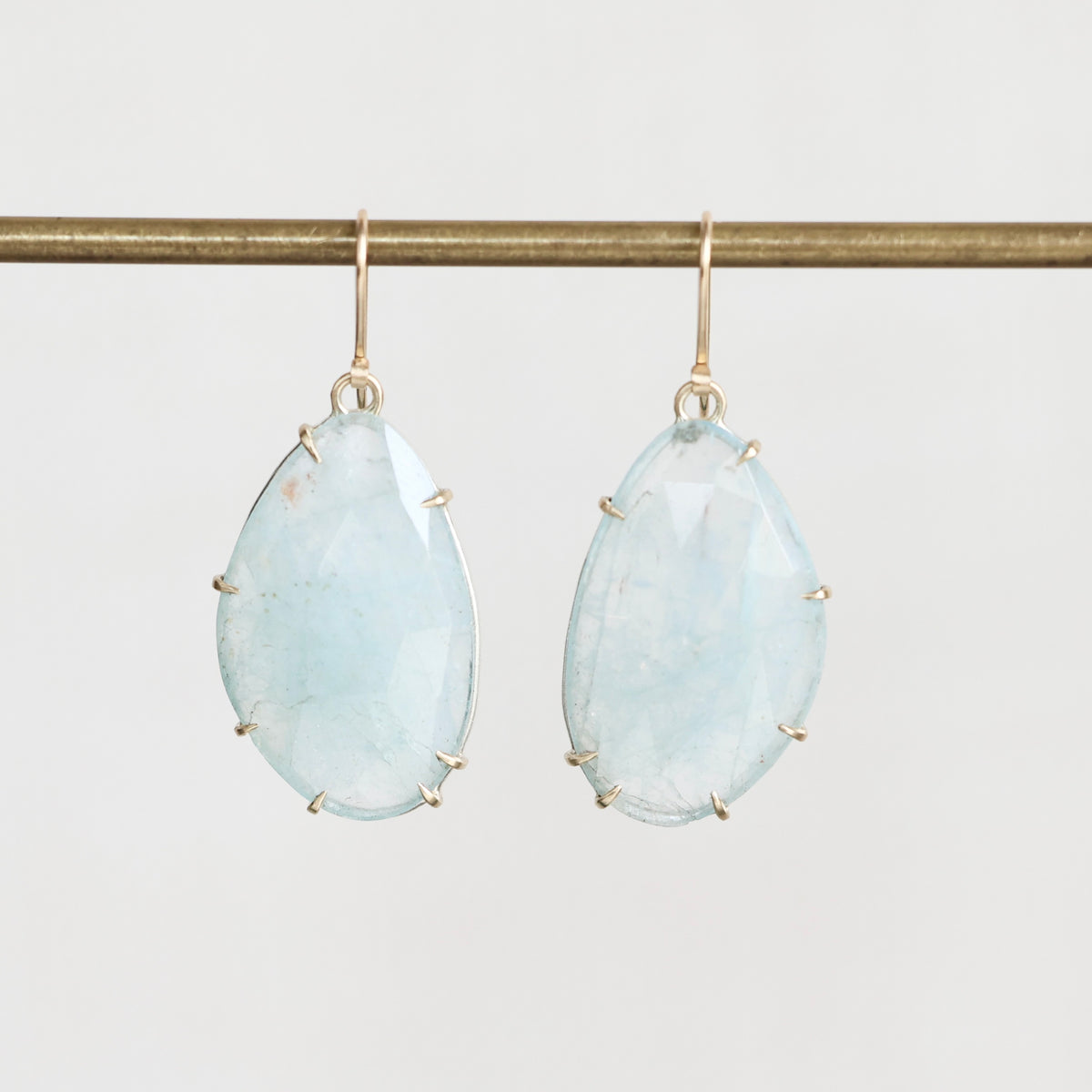 A pair of teardrop-shaped light blue gemstone drop earrings with gold hooks and prong settings, hanging from a horizontal gold display bar against a light gray background.
