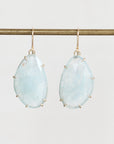 A pair of teardrop-shaped light blue gemstone drop earrings with gold hooks and prong settings, hanging from a horizontal gold display bar against a light gray background.