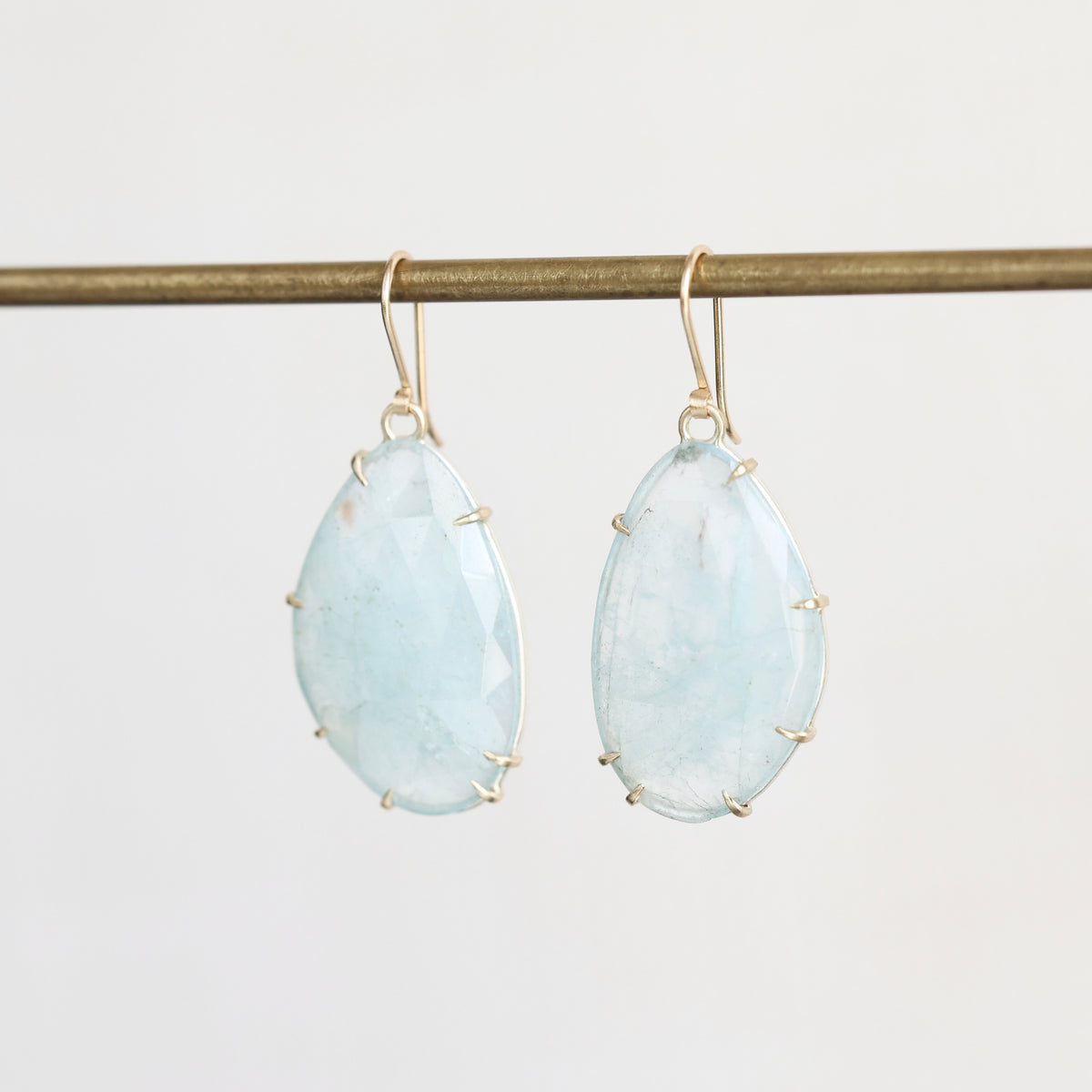 A pair of teardrop-shaped light blue gemstone drop earrings with gold hooks and prong settings, hanging from a horizontal gold rod against a plain light gray background.