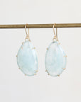 A pair of teardrop-shaped light blue gemstone drop earrings with gold hooks and prong settings, hanging from a horizontal gold rod against a plain light gray background.