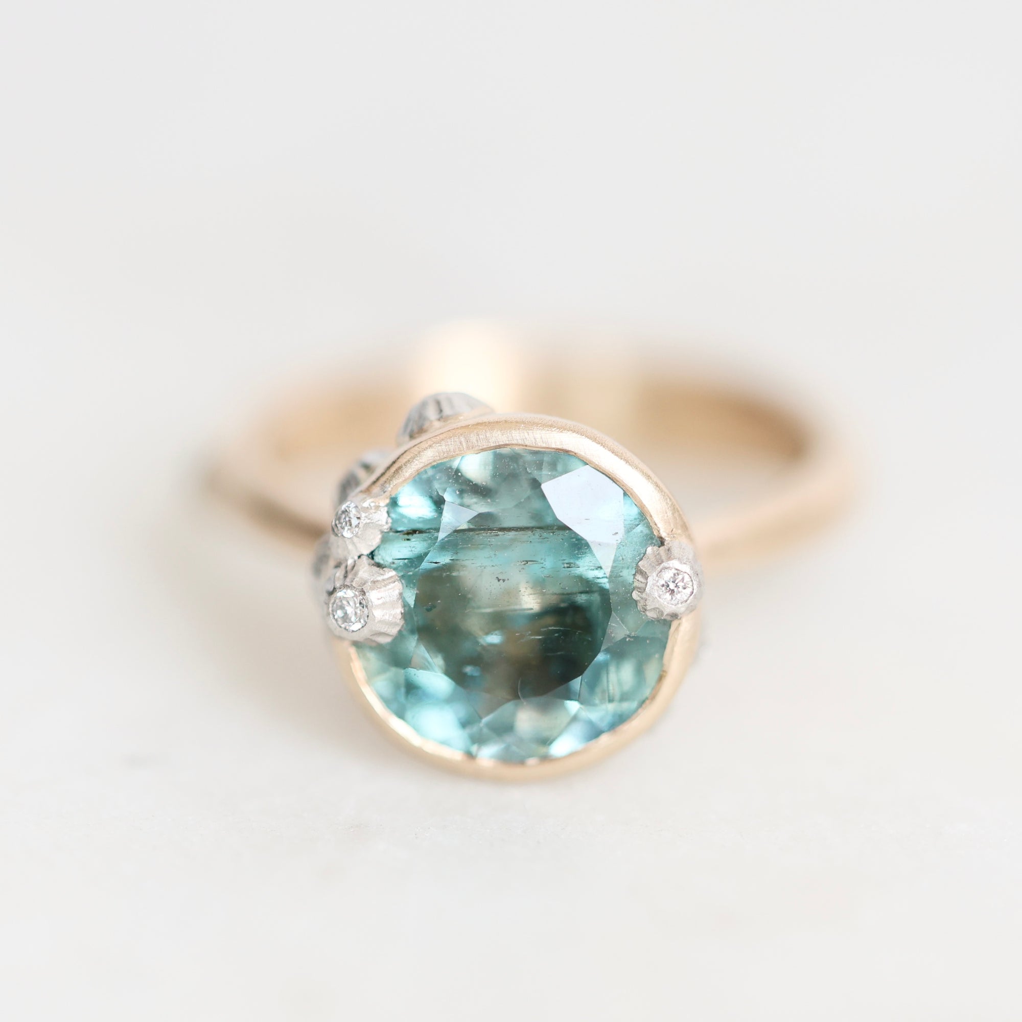 A gold ring featuring a large oval light blue gemstone in a prong setting, accented with three small round clear stones on the shoulders, photographed on a light gray background.