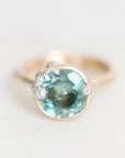 A gold ring featuring a large oval light blue gemstone in a prong setting, accented with three small round clear stones on the shoulders, photographed on a light gray background.