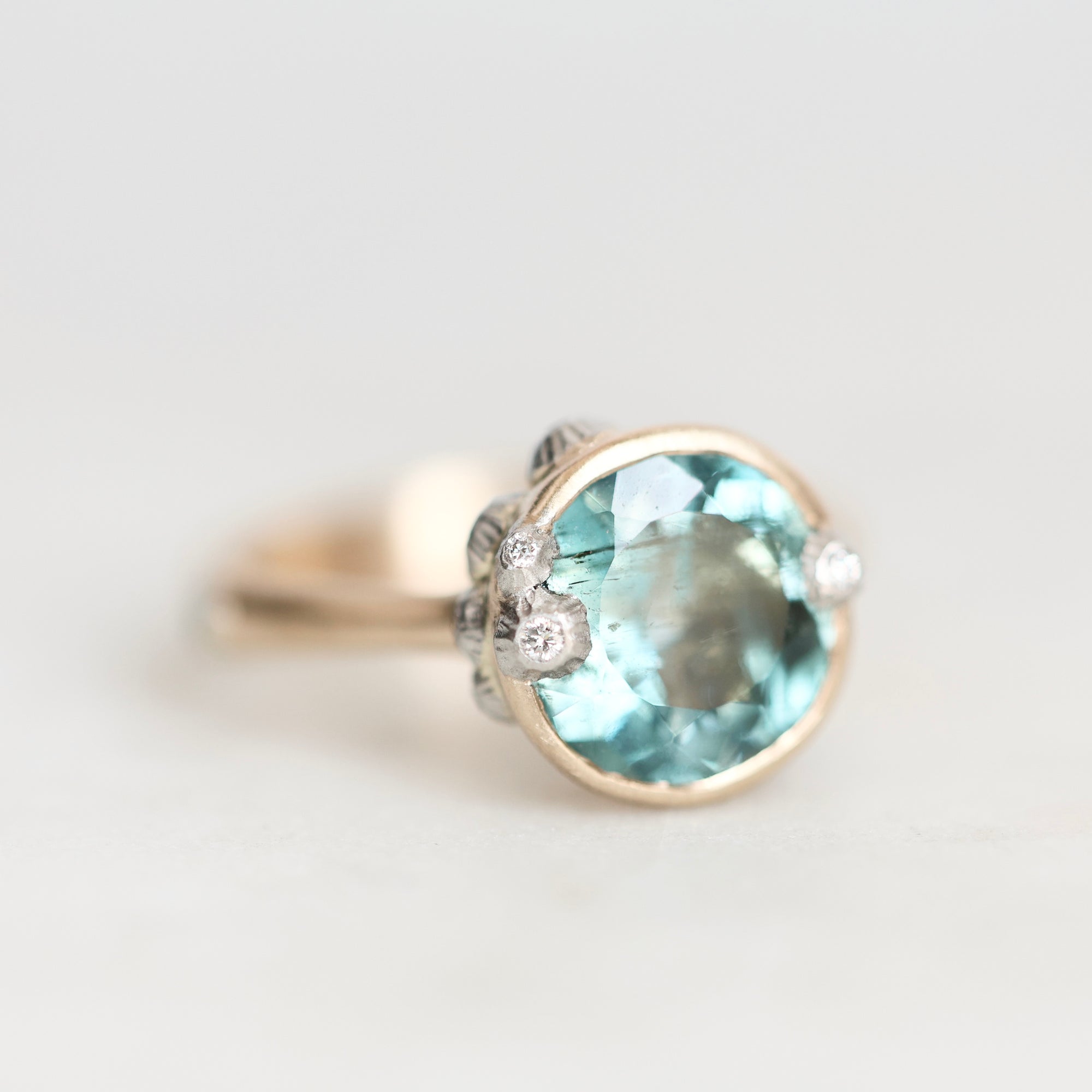 A gold ring featuring a large oval light blue gemstone in a bezel setting, accented with three small round clear stones on the band, photographed against a soft light gray background.