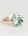 A gold ring featuring a large oval light blue gemstone in a bezel setting, accented with three small round clear stones on the band, photographed against a soft light gray background.