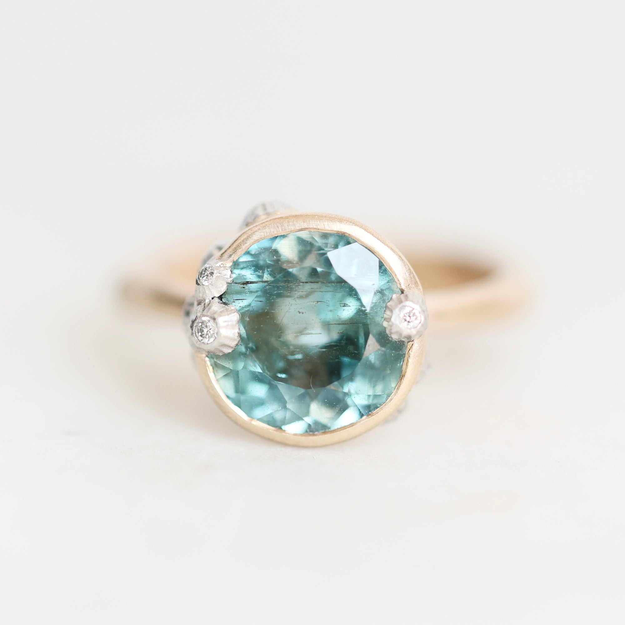 A round bezel-set ring featuring a large light blue gemstone in a gold-toned setting, accented with small clear stones on the shoulders, shown on a plain light background.