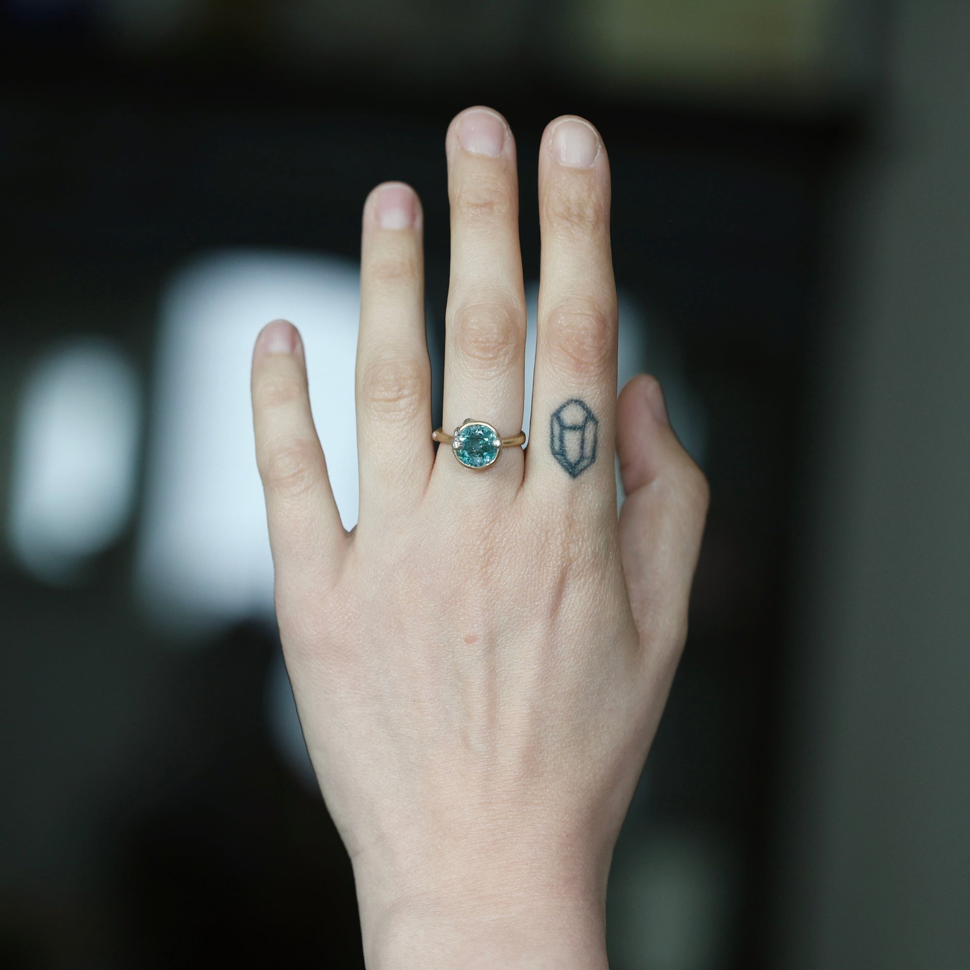 A hand wearing a gold ring with a round blue-green stone, shown against a dark blurred background. The hand also has a small geometric tattoo on the side of the index finger.